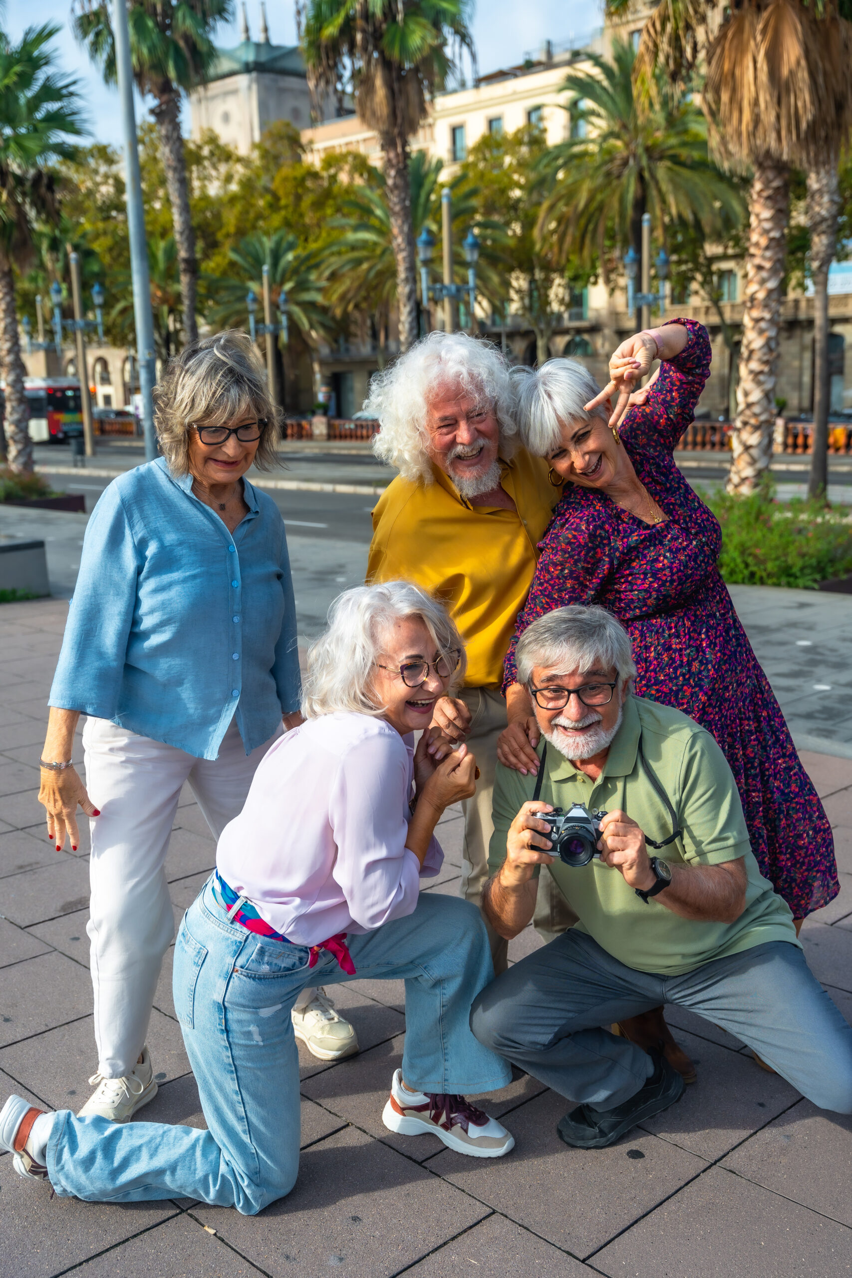Cheerful group of senior friends traveling together, capturing fun memories with an analog camera while exploring a city street lined with palm trees on a sunny day
